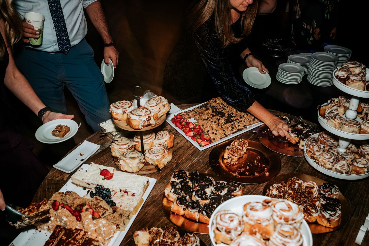 Guests gather around a rustic dessert table filled with cinnamon rolls, pastries, and sweet treats at a wedding reception.