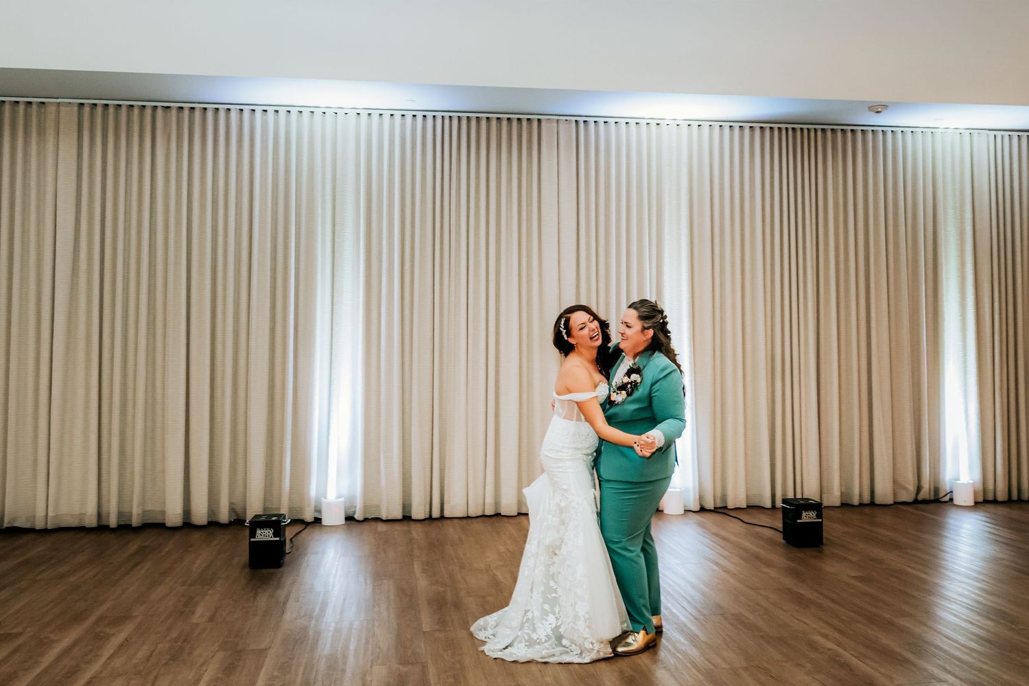 Two brides share their first dance in a teal suit and white lace gown on a wooden dance floor.
