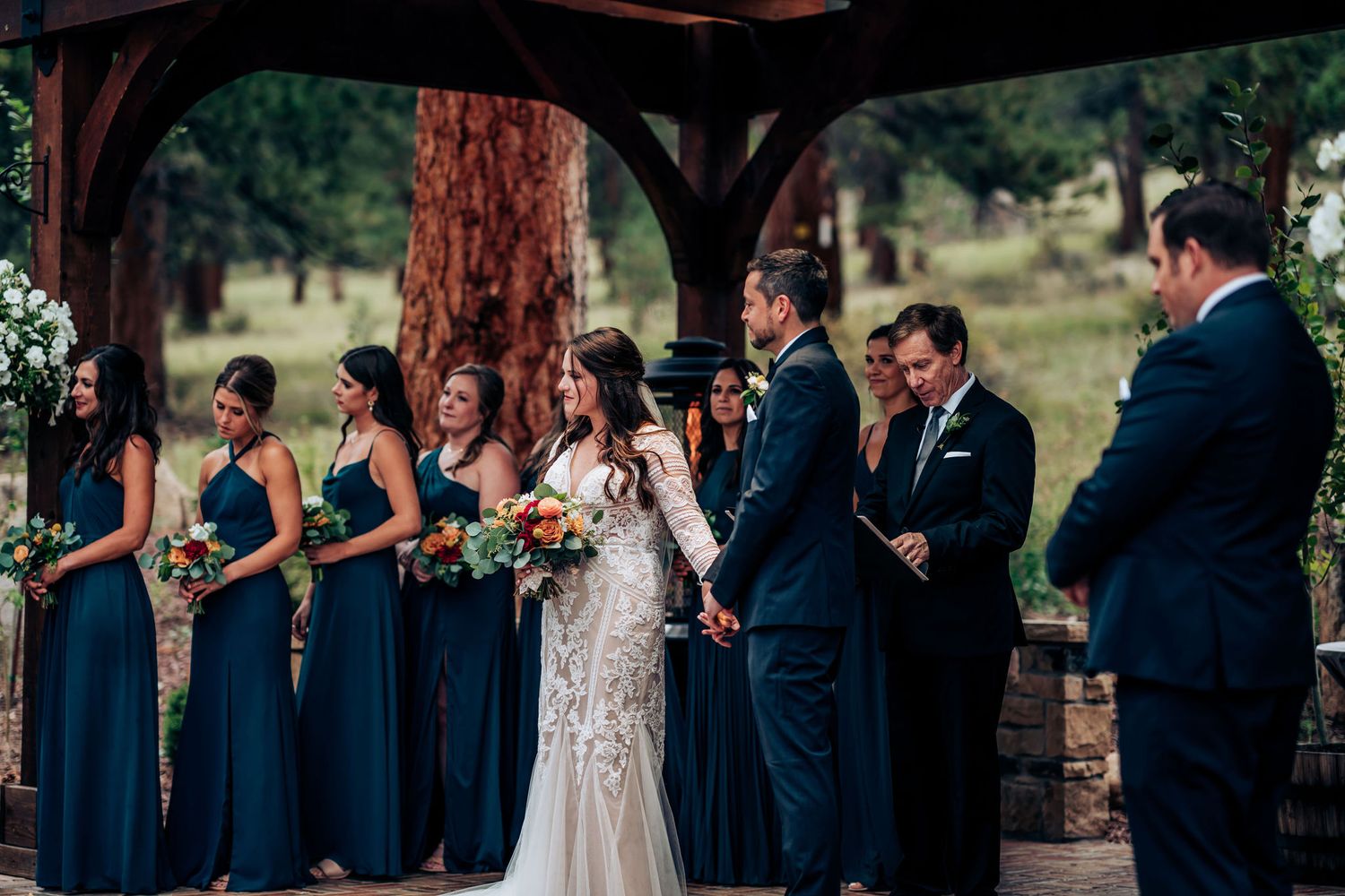 Bridesmaids in dark blue gowns line up beside the wedding couple during an outdoor ceremony.