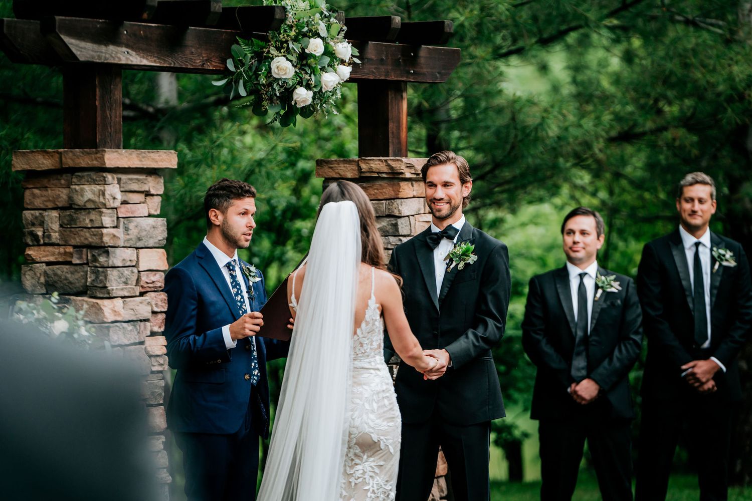 A wedding ceremony takes place under a stone archway surrounded by lush greenery.
