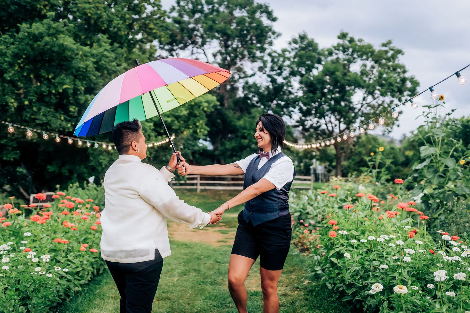 Two people holding hands and dancing with a rainbow umbrella in a flower garden.