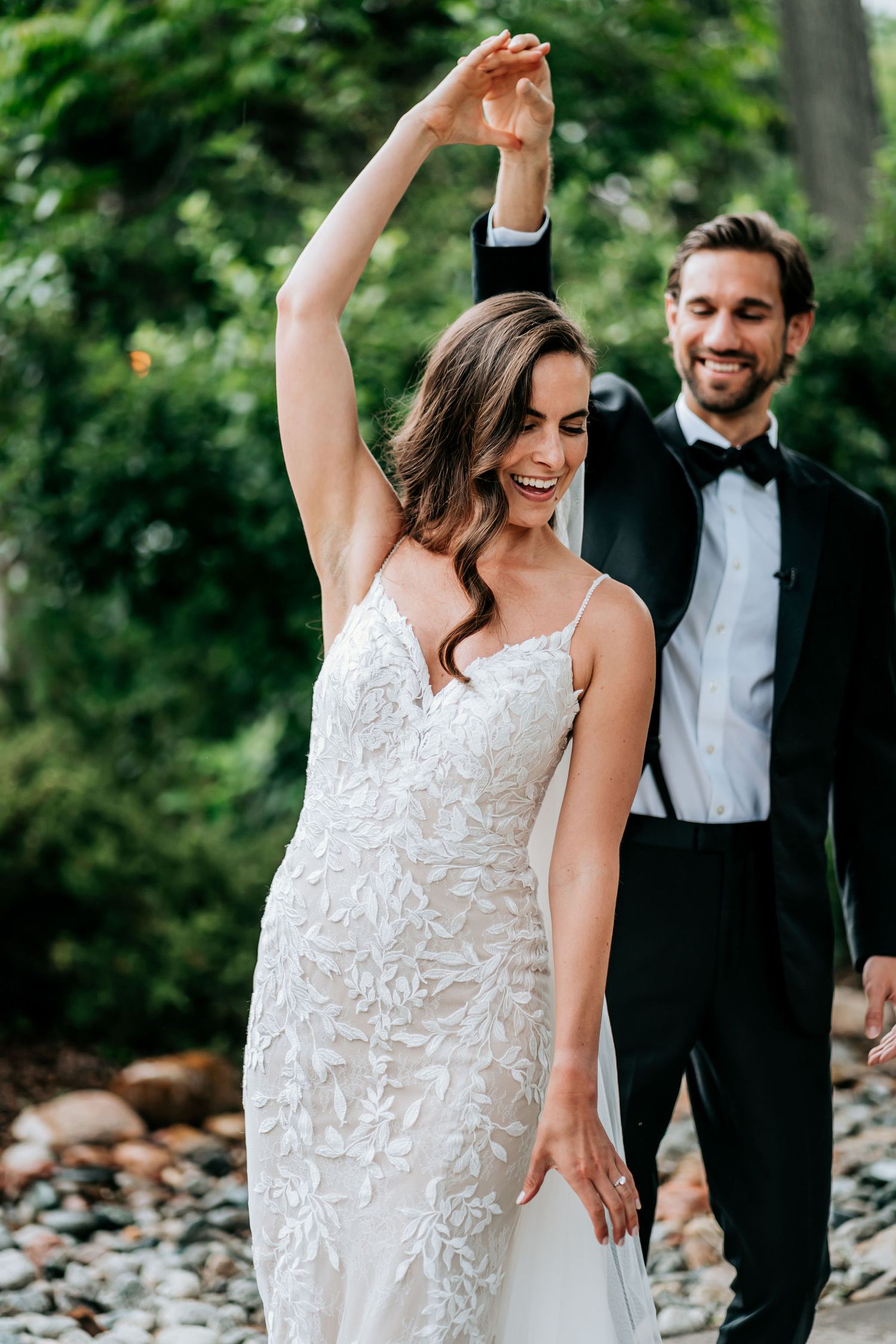 A couple in wedding attire dance joyfully outdoors with lush greenery in the background.
