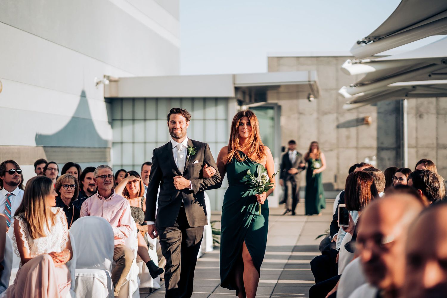 Formal procession walking down an outdoor aisle between rows of guests at a rooftop venue.