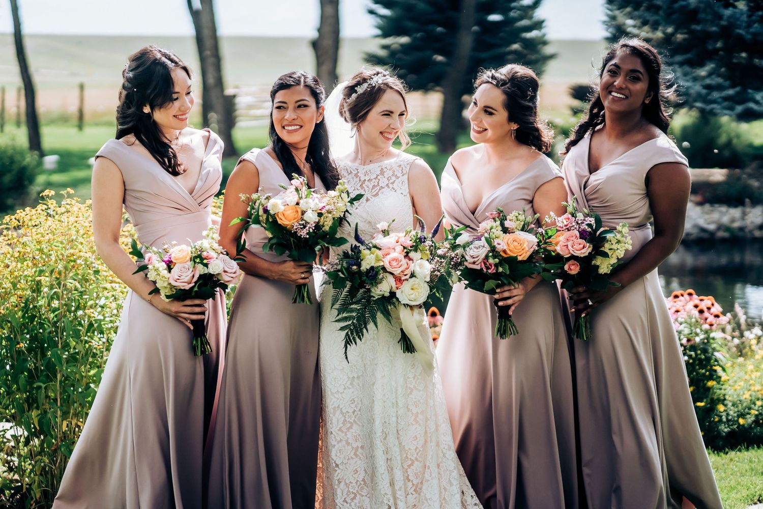 Bridal party in dusty rose dresses holding floral bouquets pose together in garden setting.