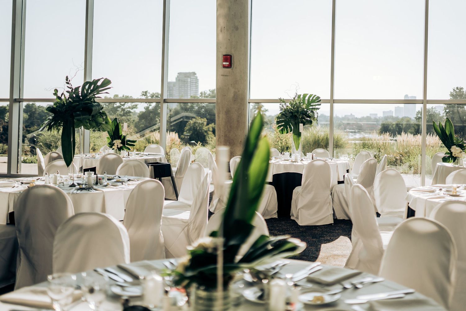 A bright wedding reception space with floor-to-ceiling windows, white-covered chairs, and tropical centerpieces.