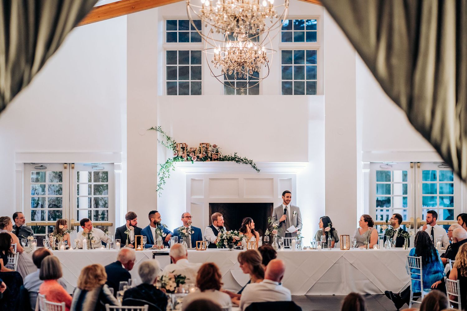 Elegant wedding reception in white room with chandelier and fireplace as guests enjoy speeches.