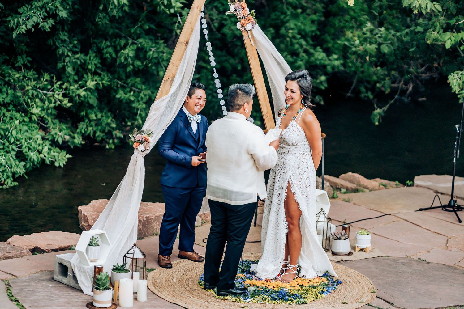 Wedding ceremony under a wooden triangle arch with white draping and floral decorations by a riverbank.