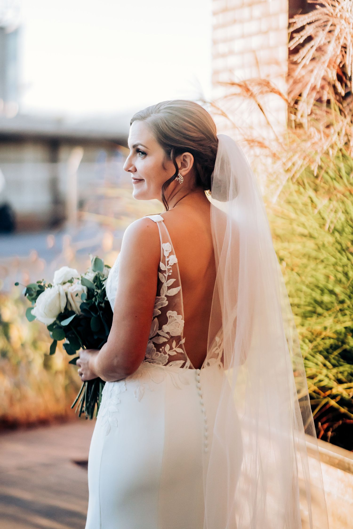 Back view of bride in elegant white dress holding bouquet against sunset light.