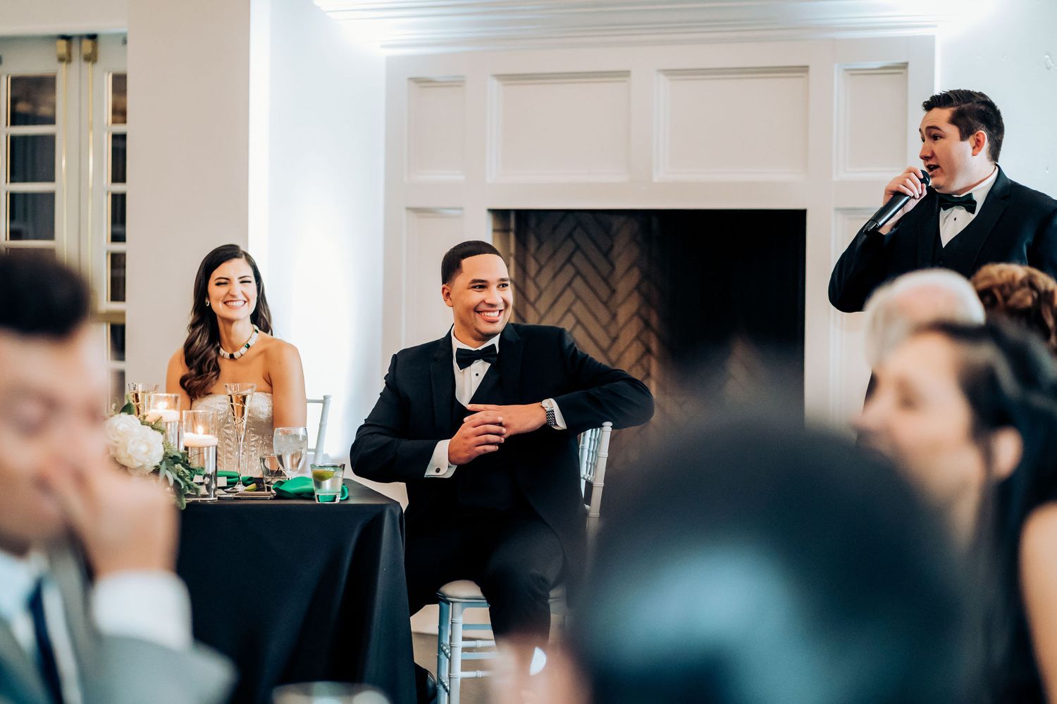 Wedding reception speech moment at head table with elegant white fireplace backdrop.
