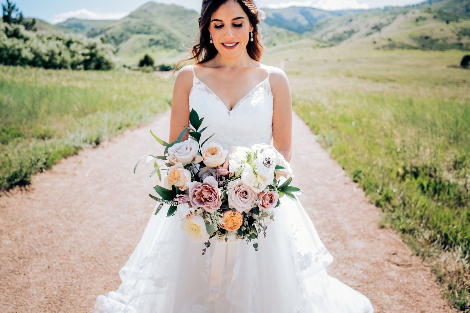 A bride in a flowing white gown holds a beautiful bouquet on a mountain dirt road.