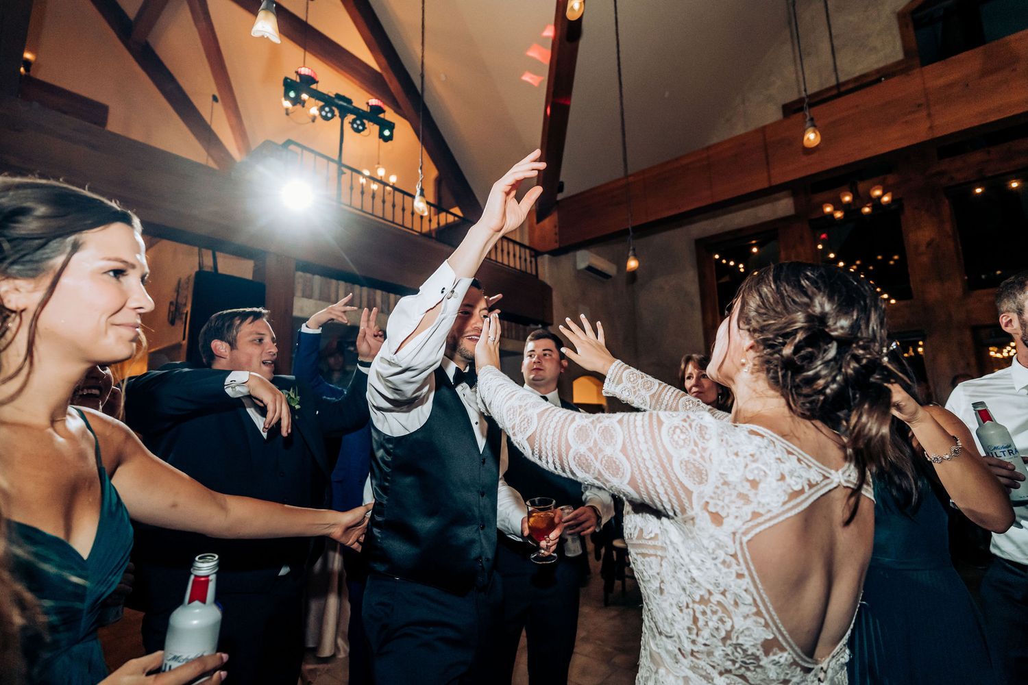Wedding guests dancing and celebrating with drinks in a rustic venue with exposed wooden beams and string lights.
