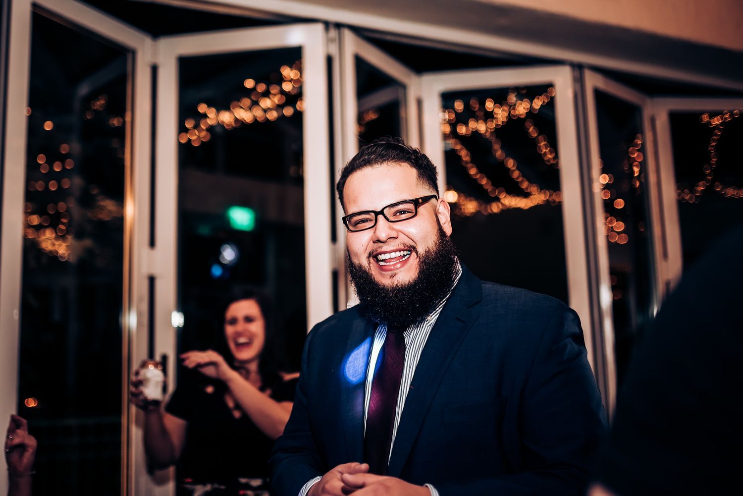 A guest in a navy suit and glasses smiles warmly at a formal indoor event.