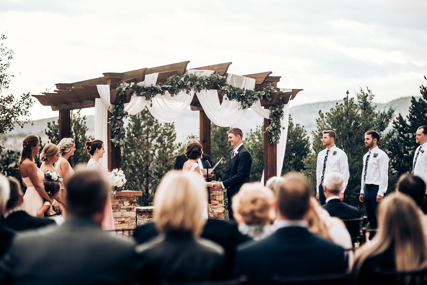 Scenic outdoor wedding ceremony with guests watching under a decorated wooden pergola.