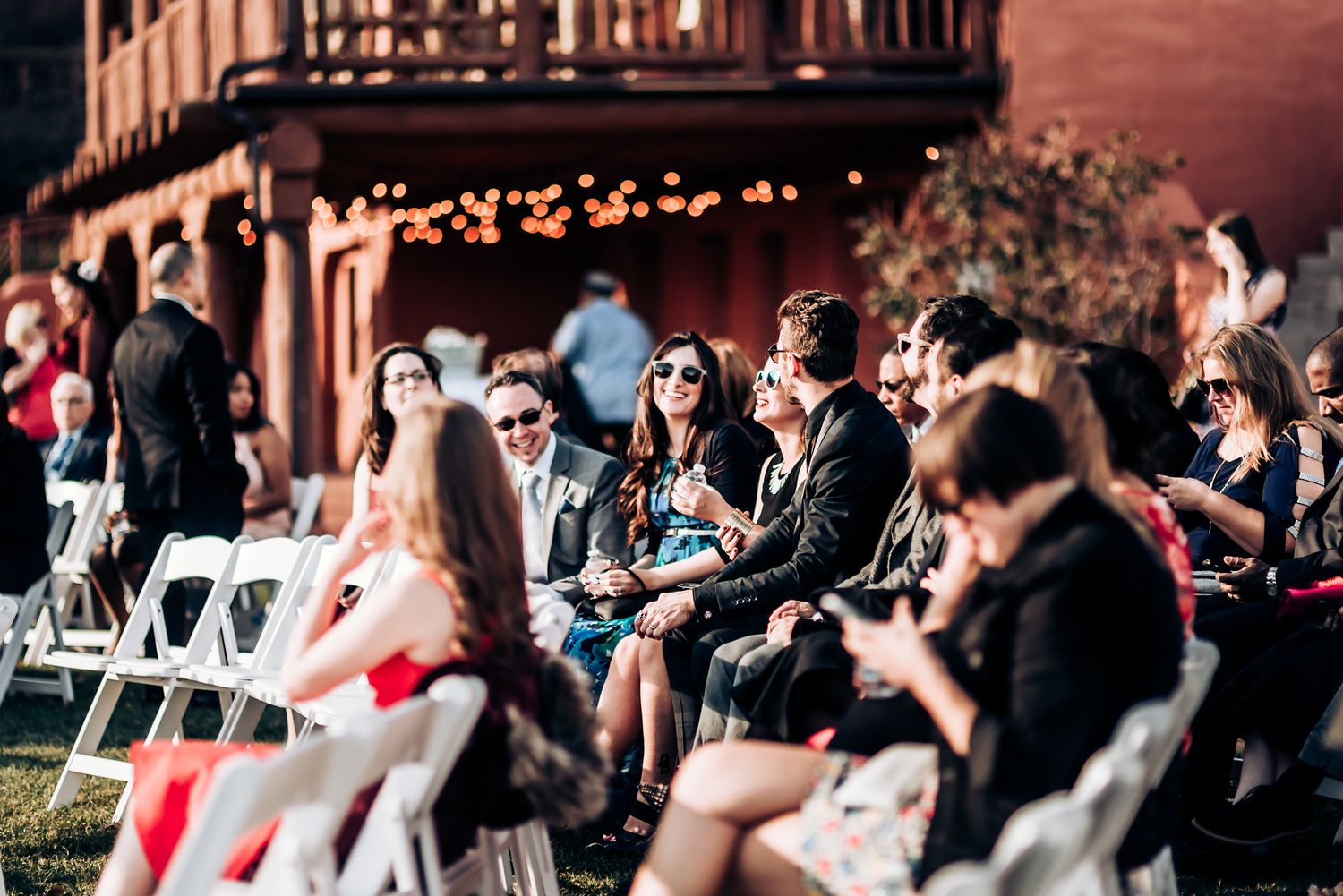 Guests seated in white chairs during an outdoor wedding ceremony with rustic building in background.