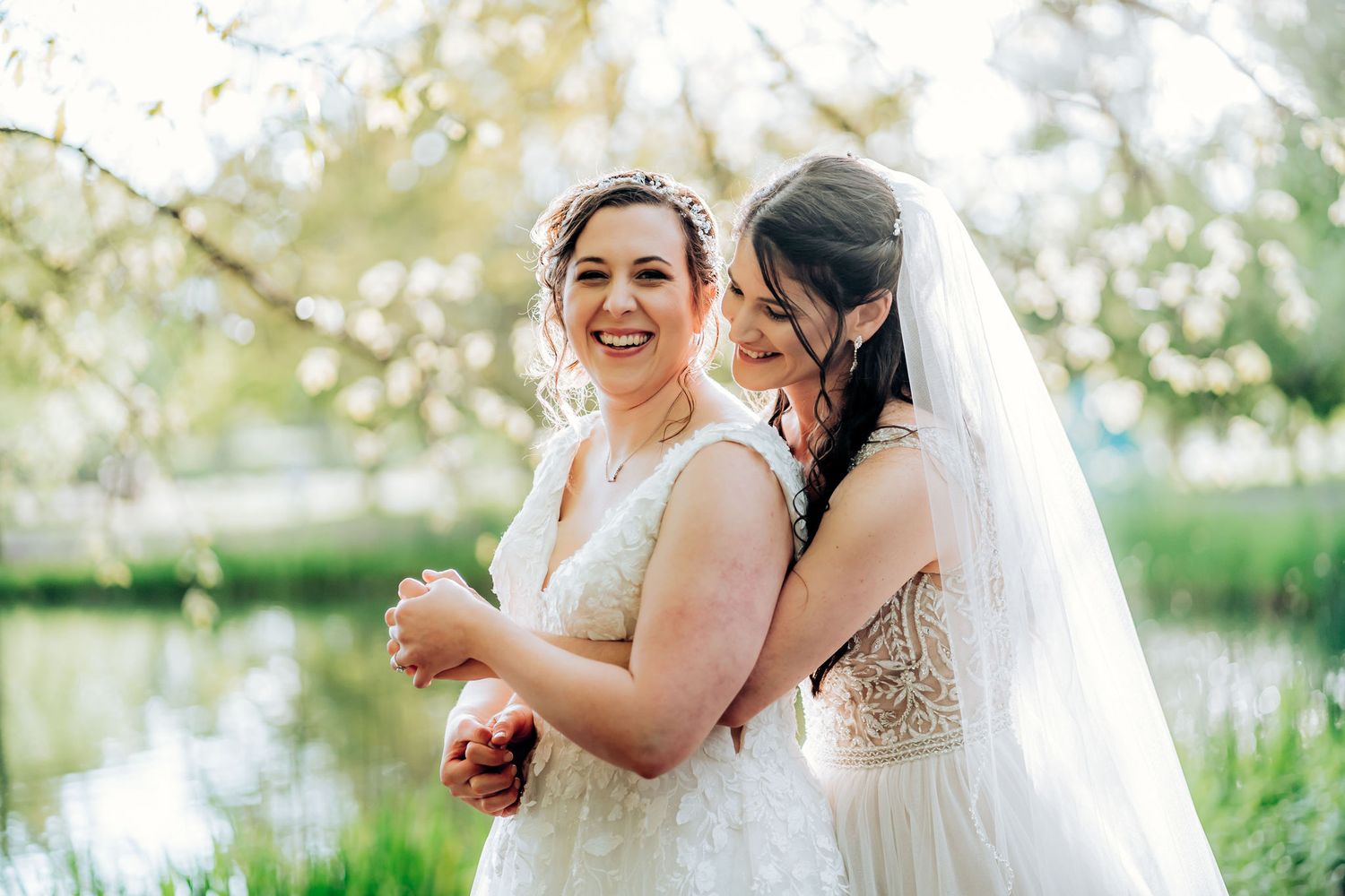 Two brides in elegant white dresses share a joyful moment together by a pond.