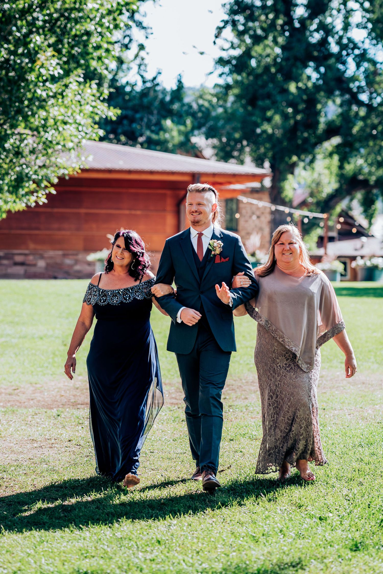 Wedding party members walk across sunny lawn with elegant formal attire at outdoor ceremony.