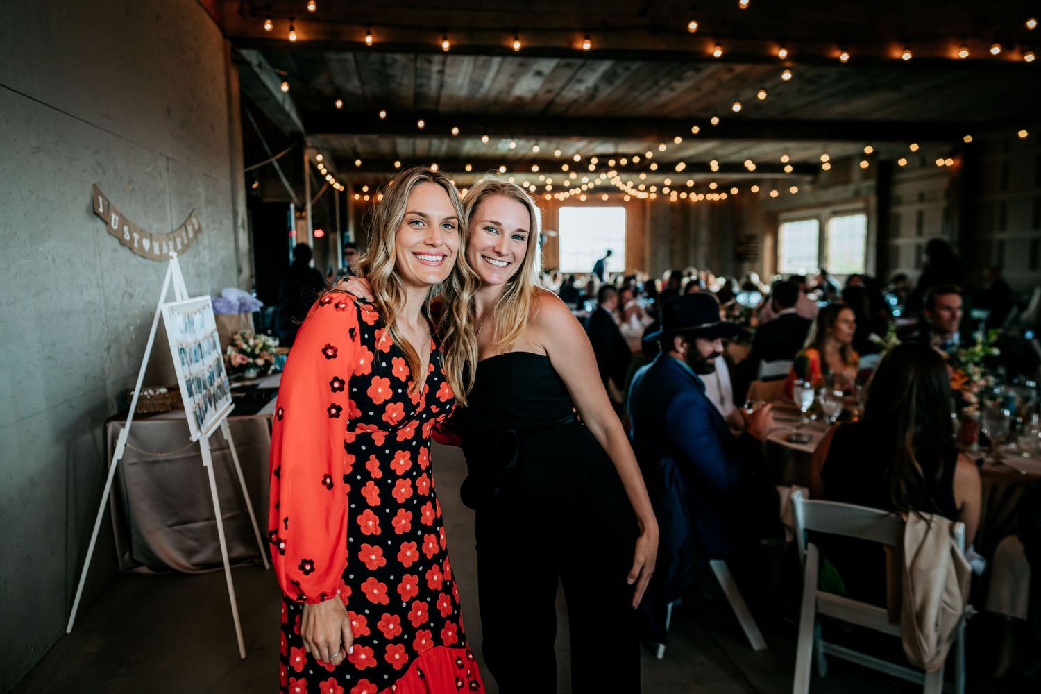 Two guests pose together at an indoor wedding reception with string lights and guests in the background.