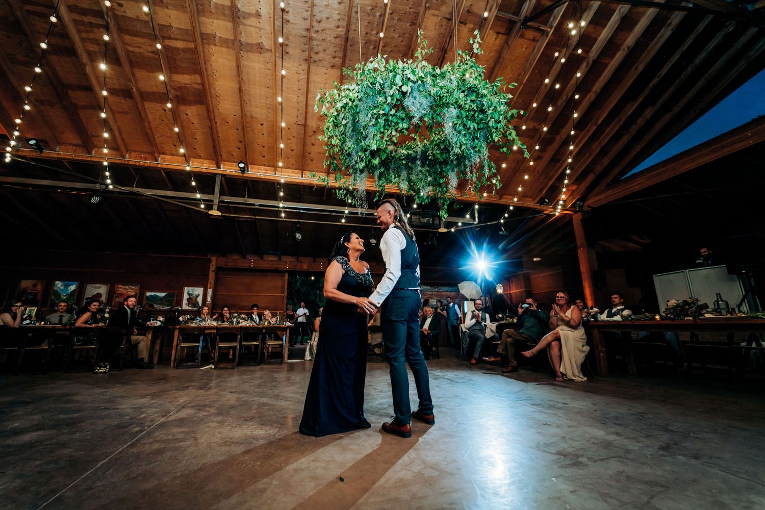 A couple shares their first dance under a hanging greenery installation in a rustic venue.