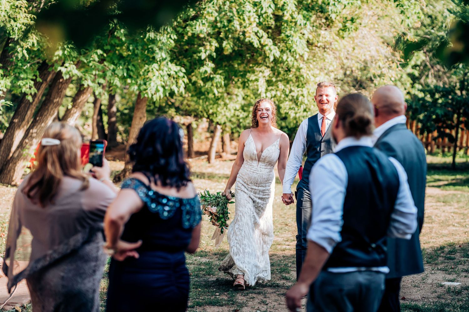 Wedding guests gather outdoors under trees for a celebration in a natural setting.