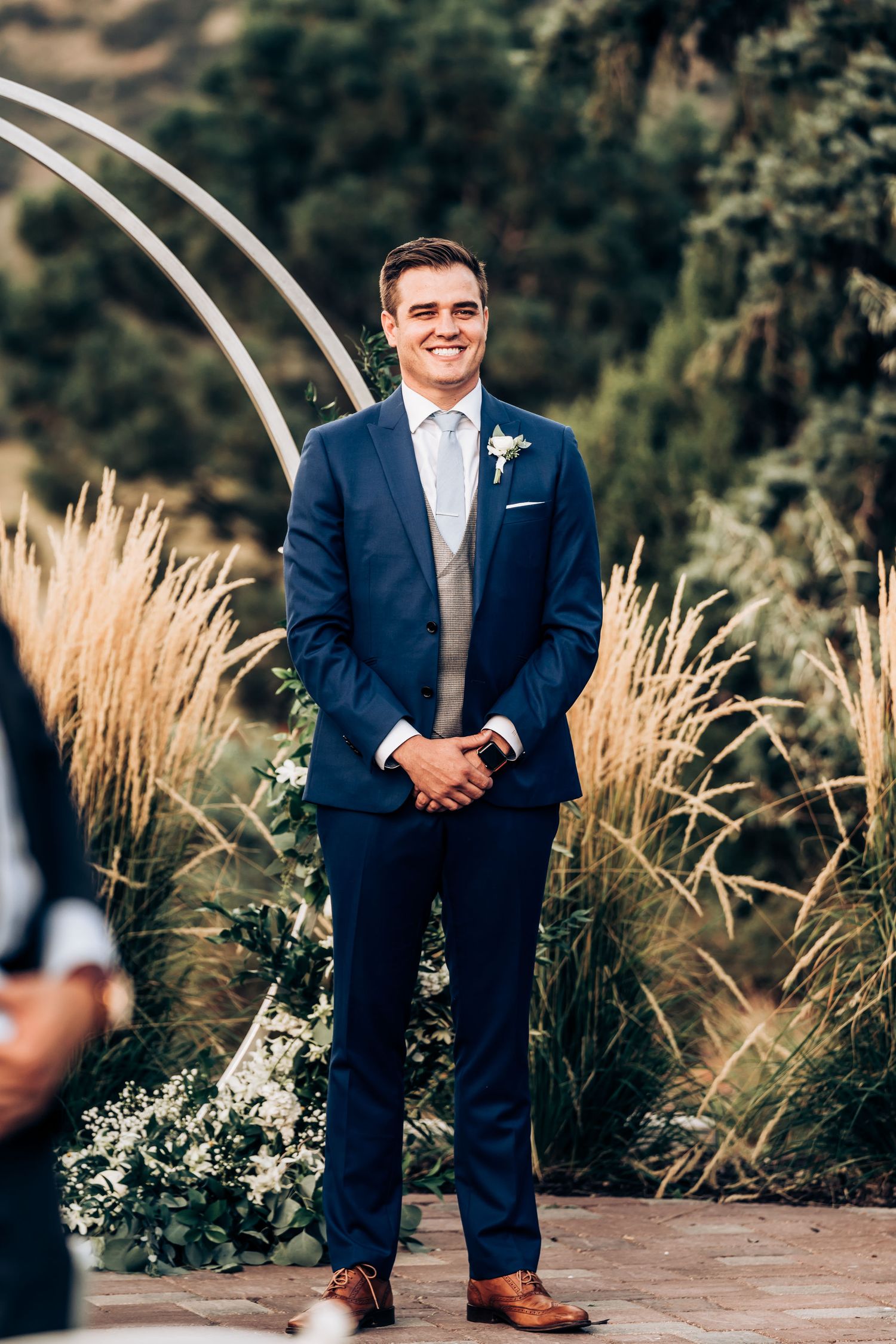 Wedding attendant in blue suit stands at altar with ornamental grasses and curved arch.