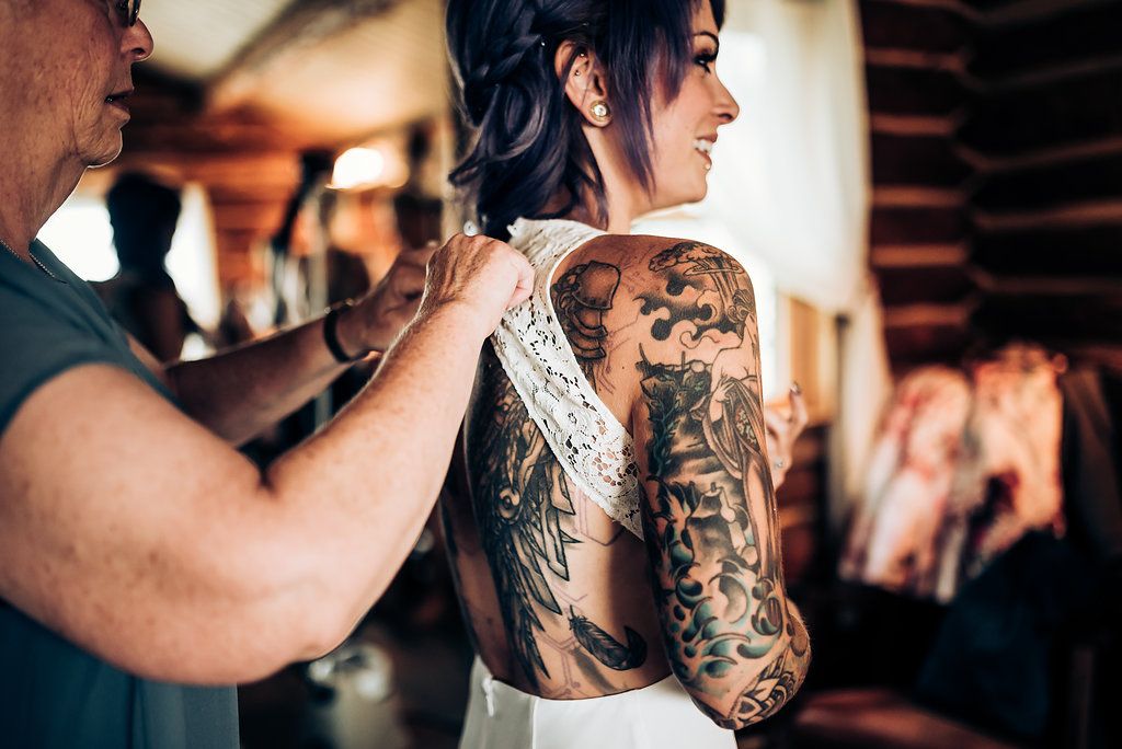 Detailed back tattoo visible through open-back white wedding dress during preparation.