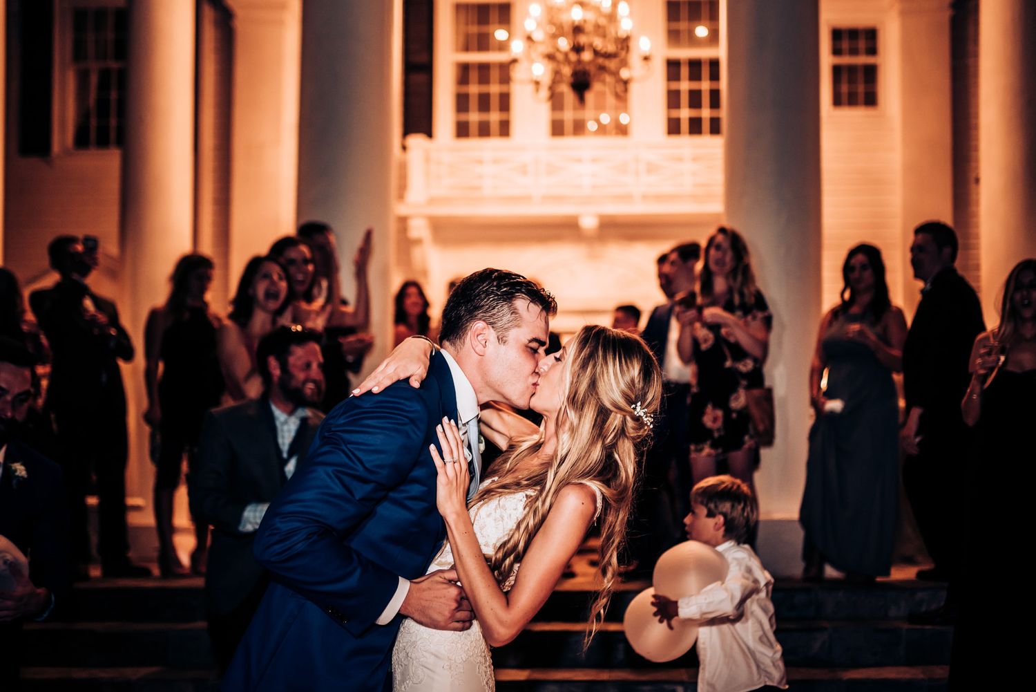 A romantic evening kiss between a couple on an elegant columned porch with onlookers in the background at sunset.