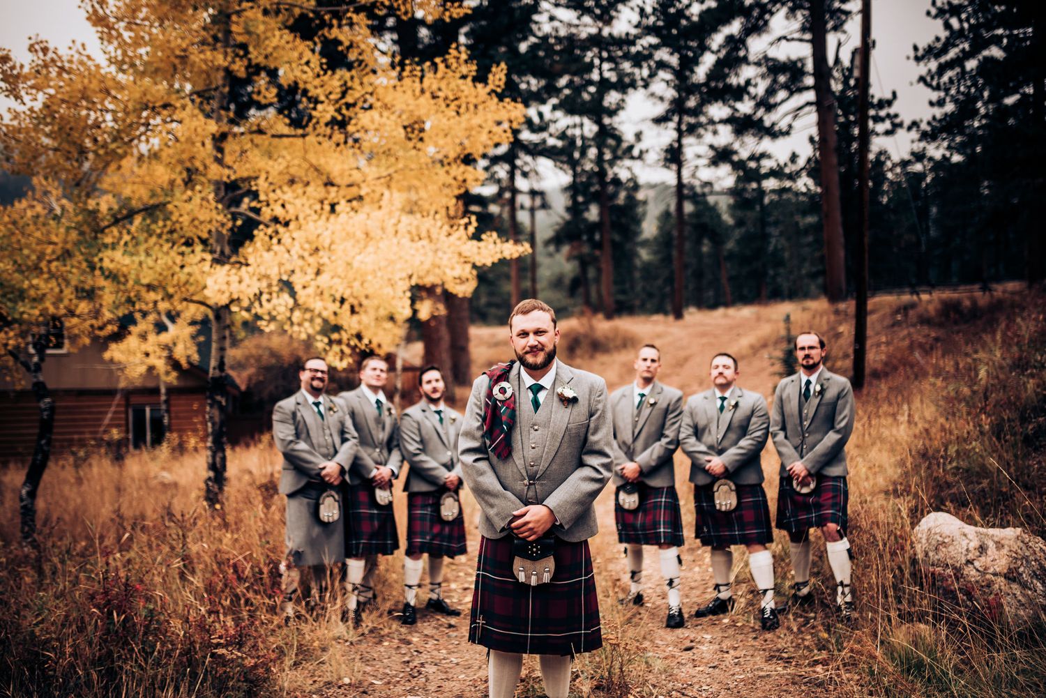 Groomsmen in traditional Scottish kilts stand in formation on outdoor path.