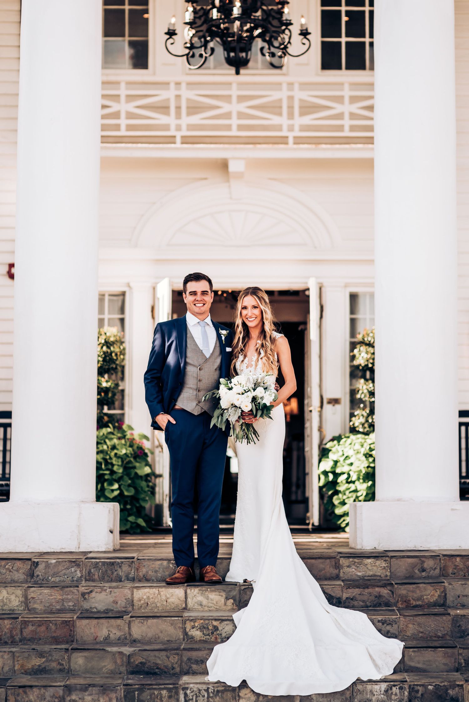 Wedding couple standing on stone steps in front of elegant white pillared venue entrance.
