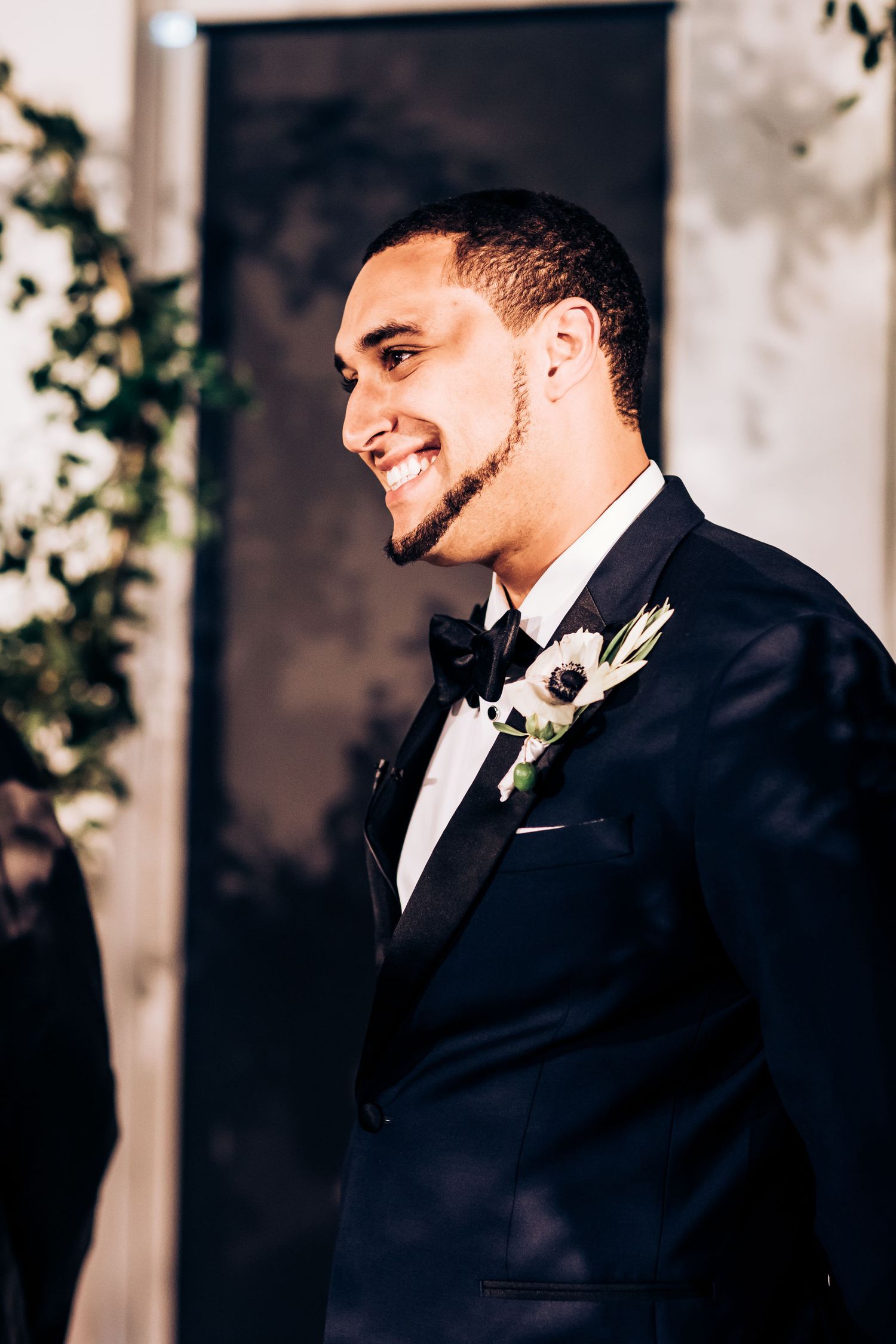 A groom in a classic black tuxedo smiles during his wedding ceremony.