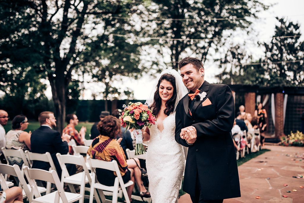 Joyful wedding recessional along outdoor path with guests seated in white chairs.
