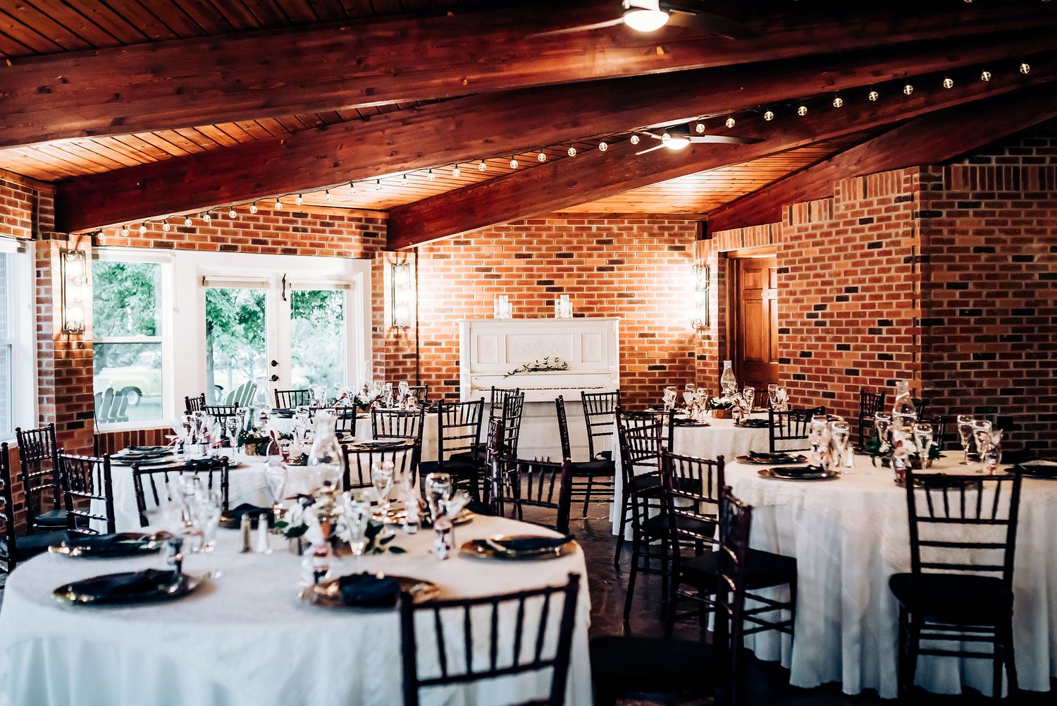 Elegant wedding reception venue with white tablecloths and dark Chiavari chairs in a rustic brick room.