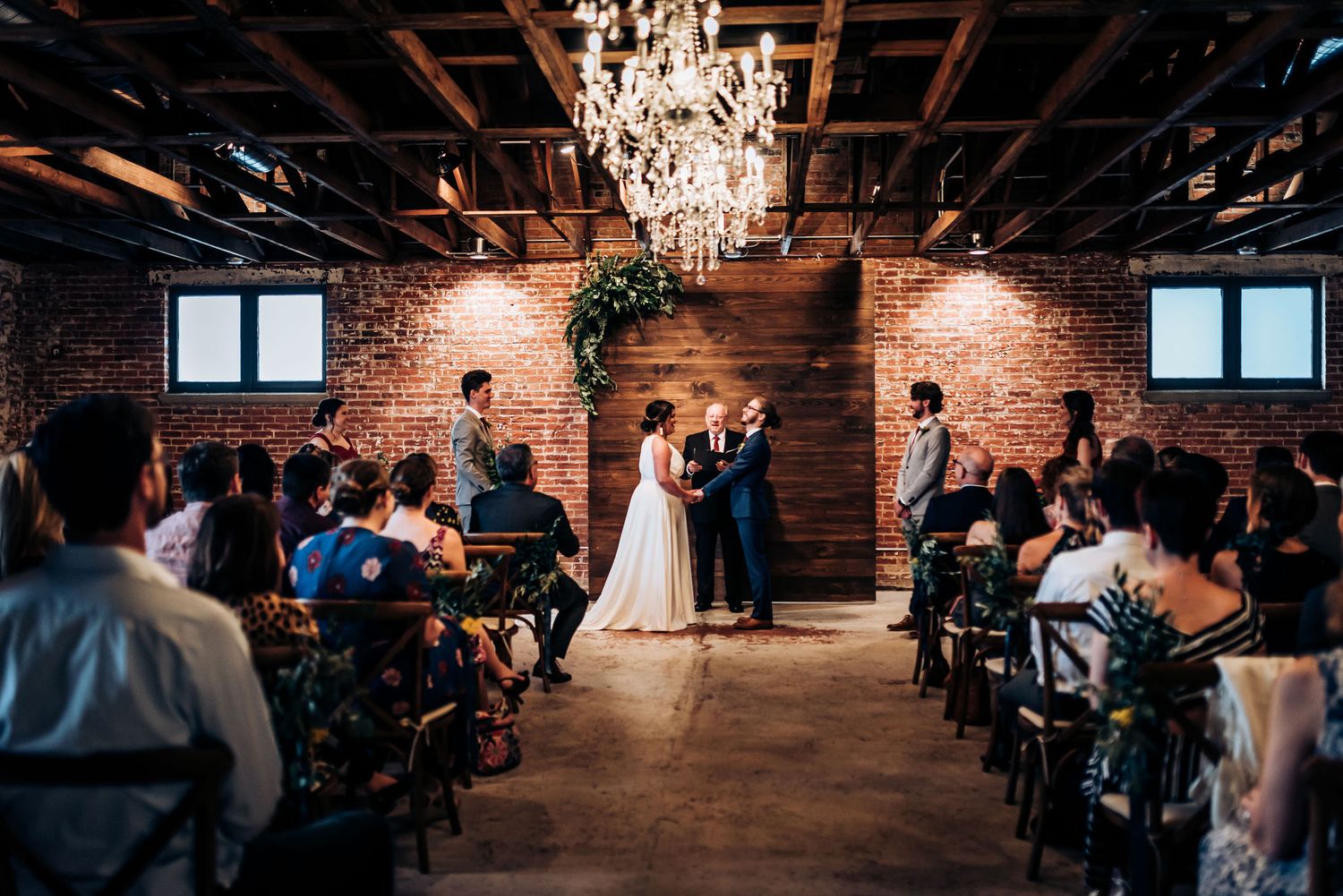 Wedding ceremony in industrial venue with chandelier and exposed brick featuring guests seated in rows.