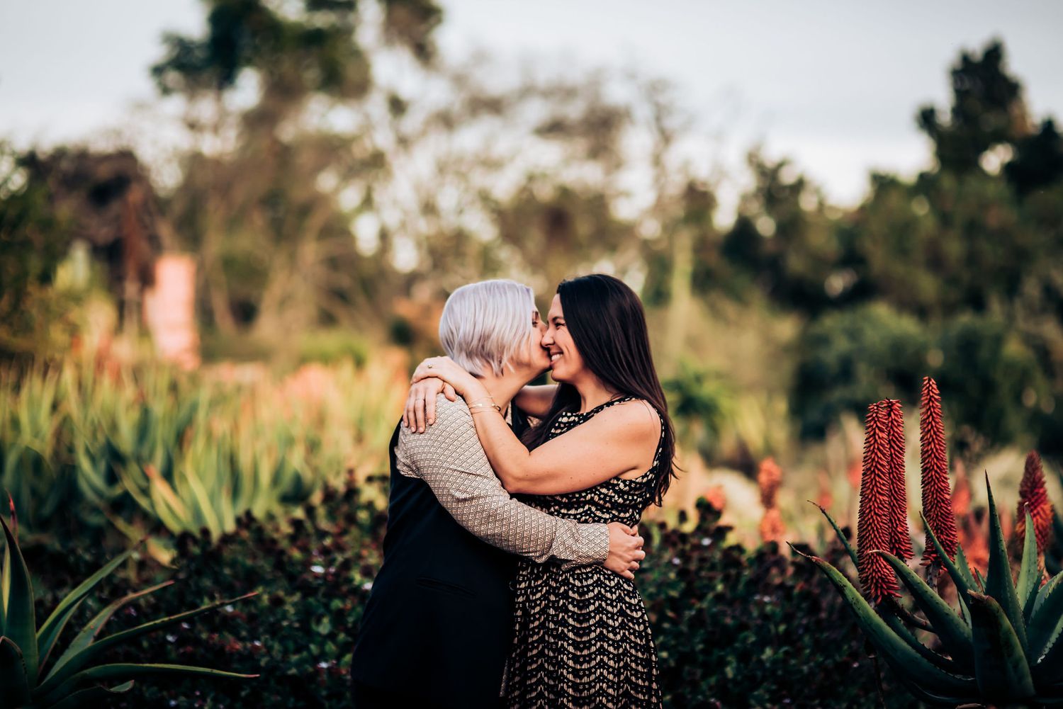 A romantic embrace in a lush garden setting with red flowers and greenery at sunset.