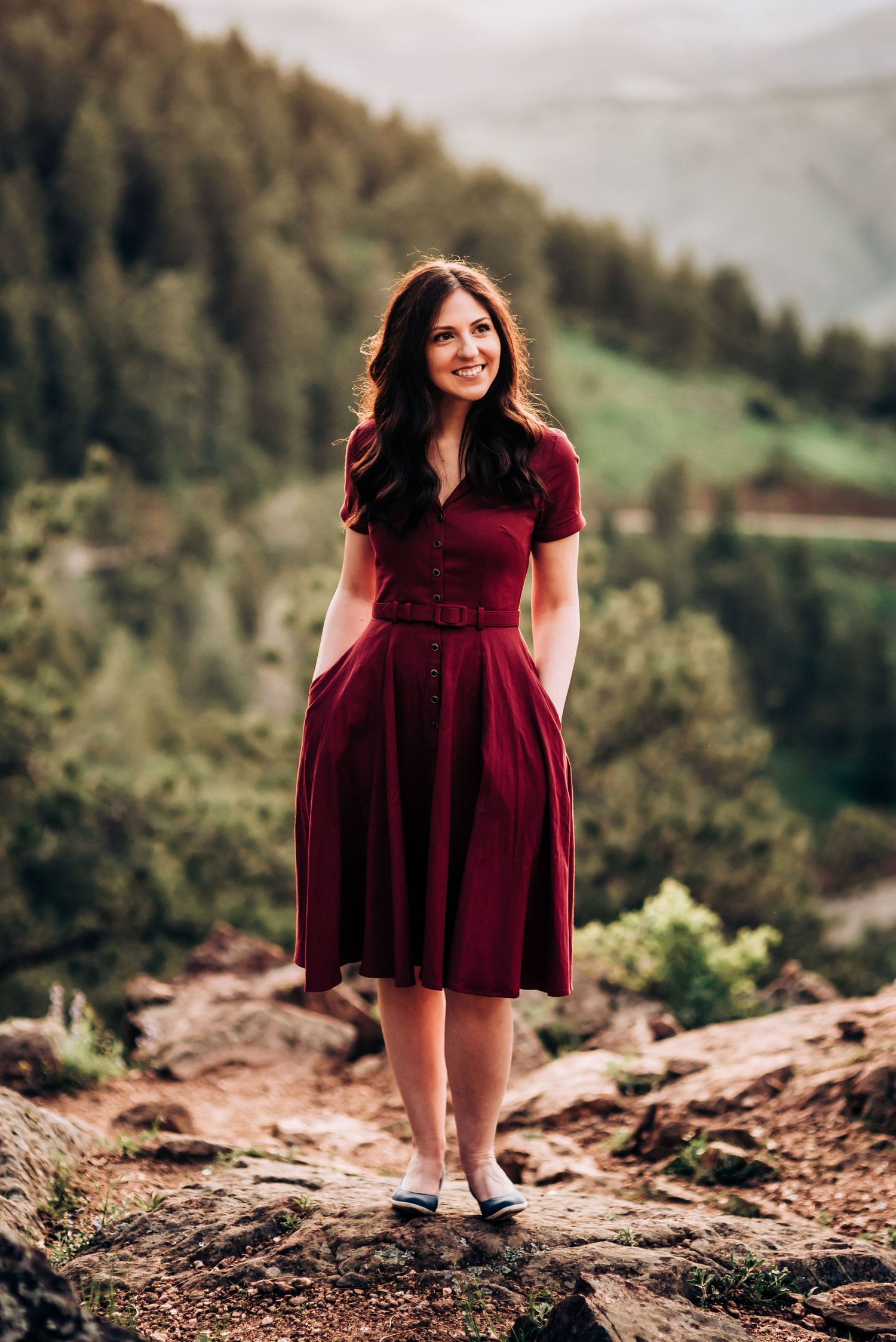 Woman in burgundy dress stands on rocky mountain overlook with scenic valley view.