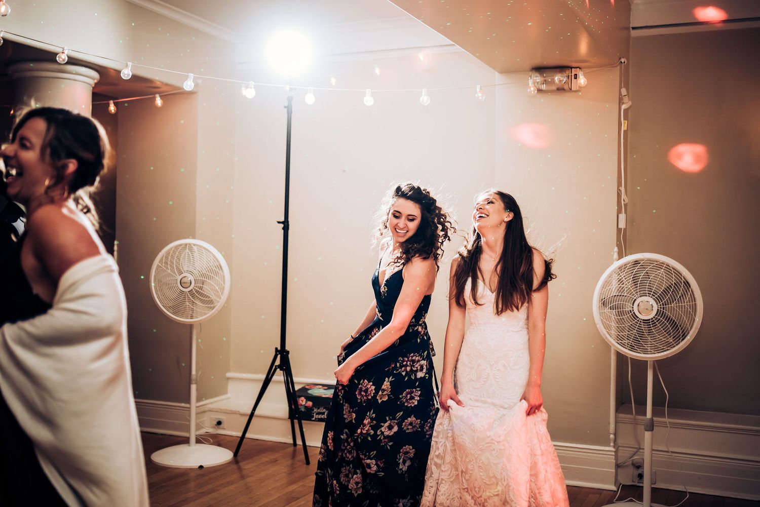 Two party guests enjoy themselves near standing fans at an indoor celebration.