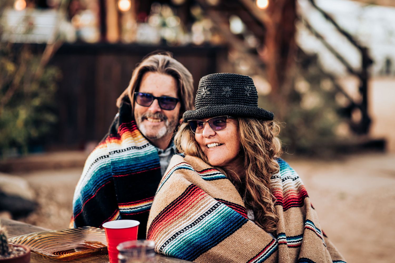 Couple wrapped in colorful serape blankets enjoying drinks outdoors.