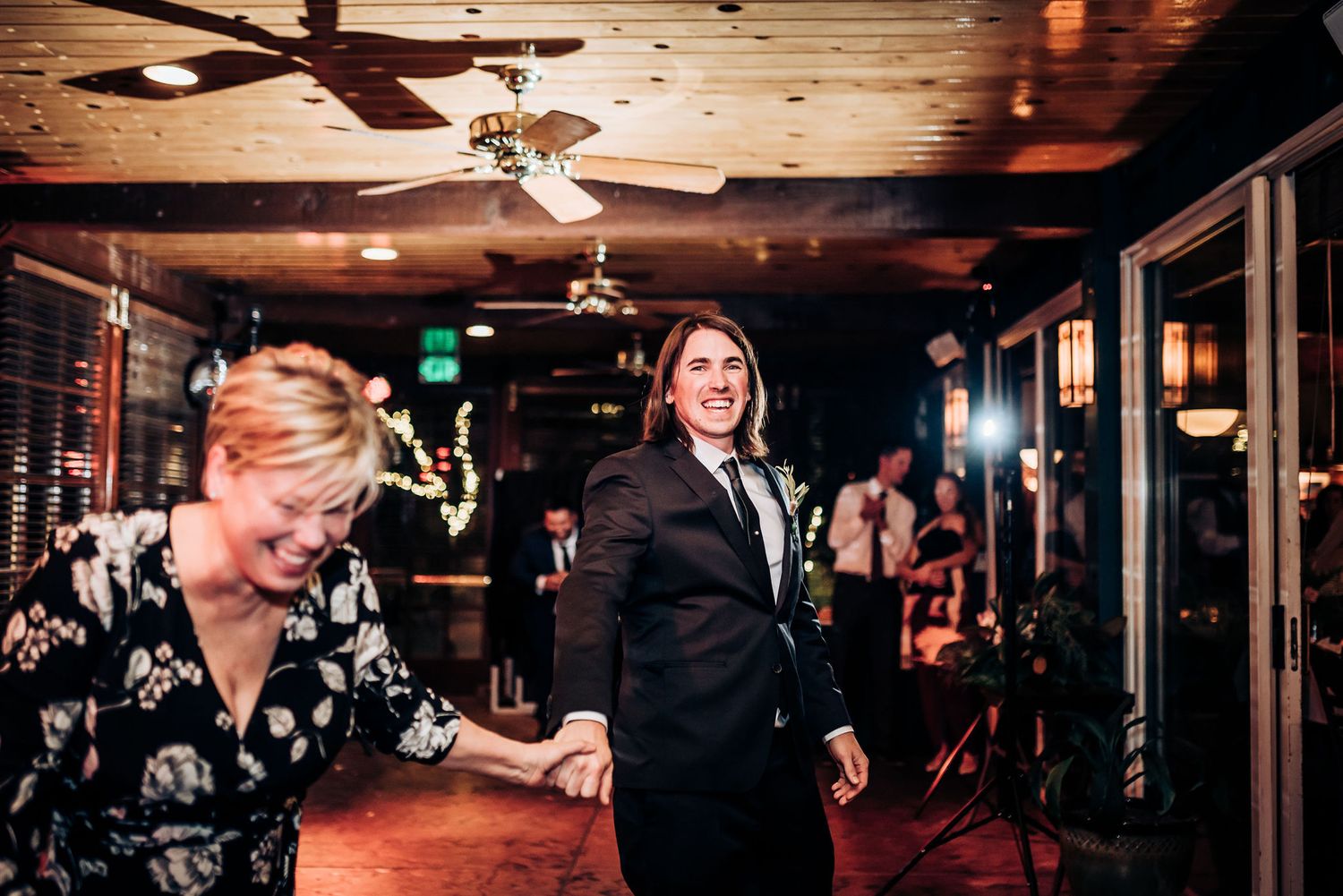 People dancing and celebrating at an indoor reception with wooden ceiling and ceiling fans.