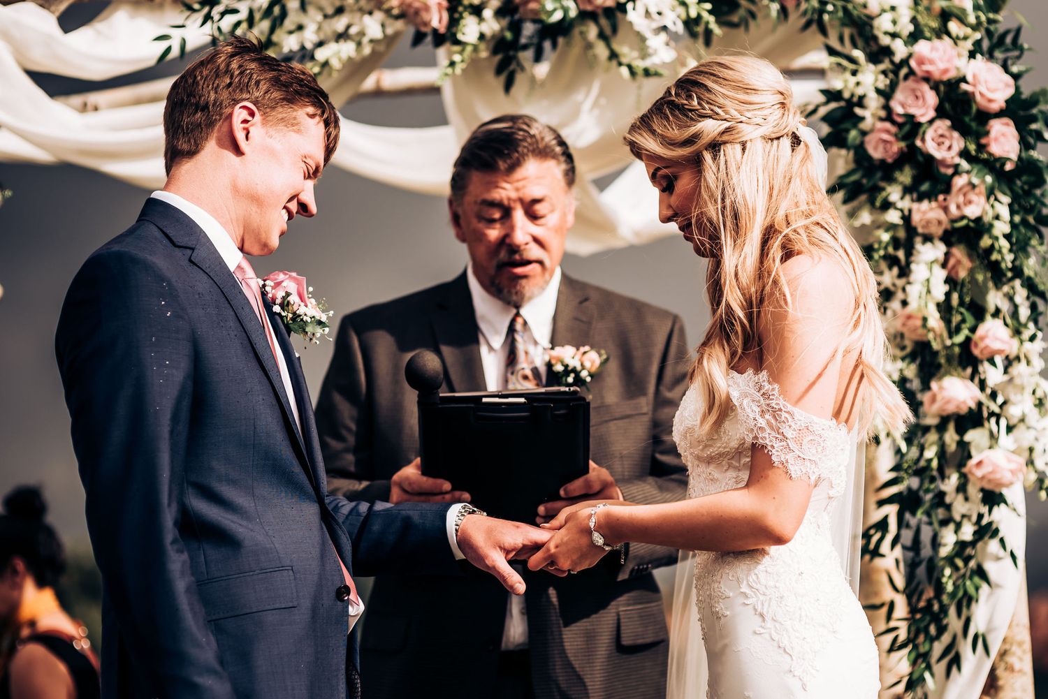 Wedding ceremony moment under floral arch as couple exchanges rings with officiant.