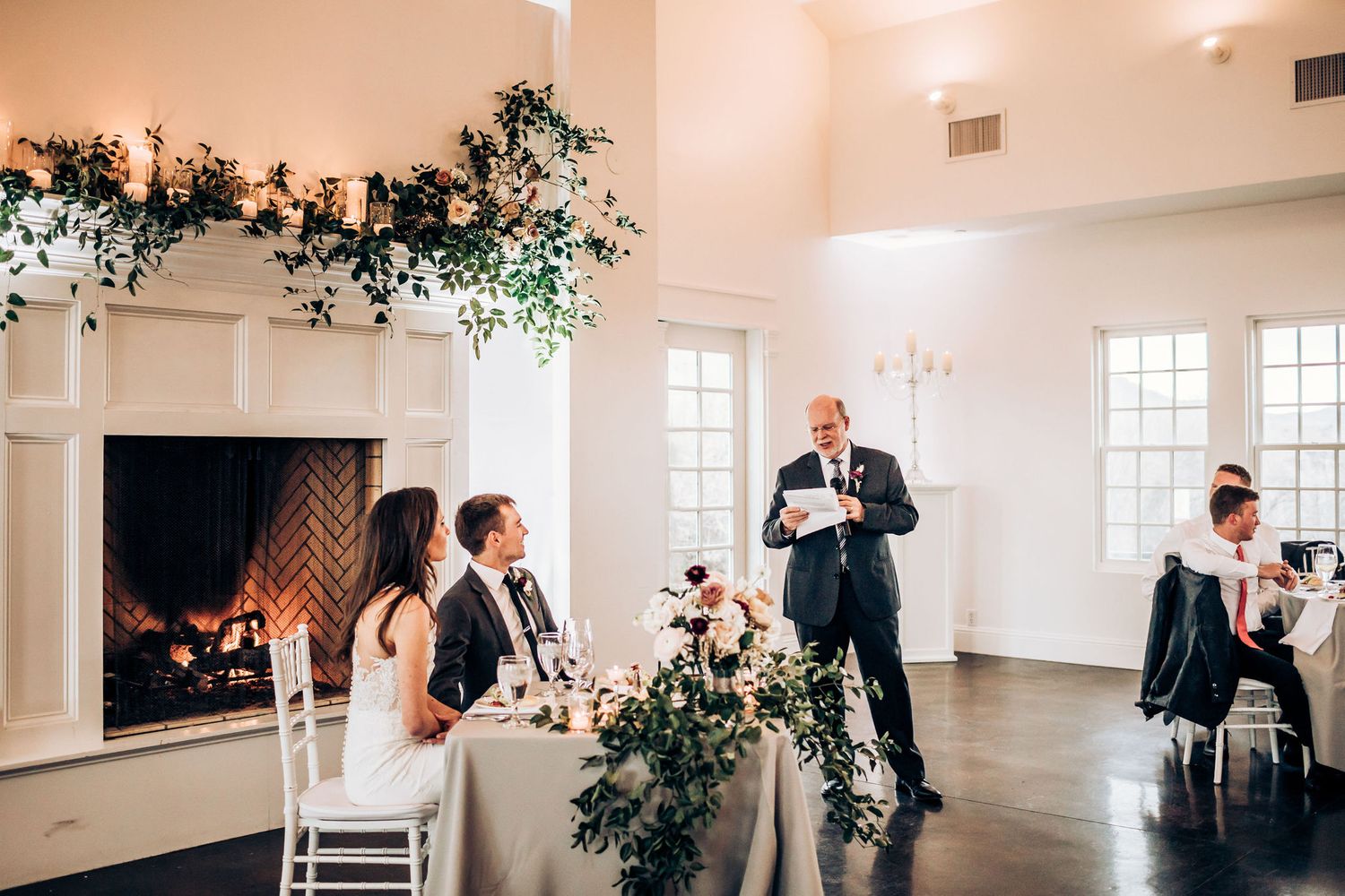 Wedding reception scene with fireplace and floral decorations during toasts.