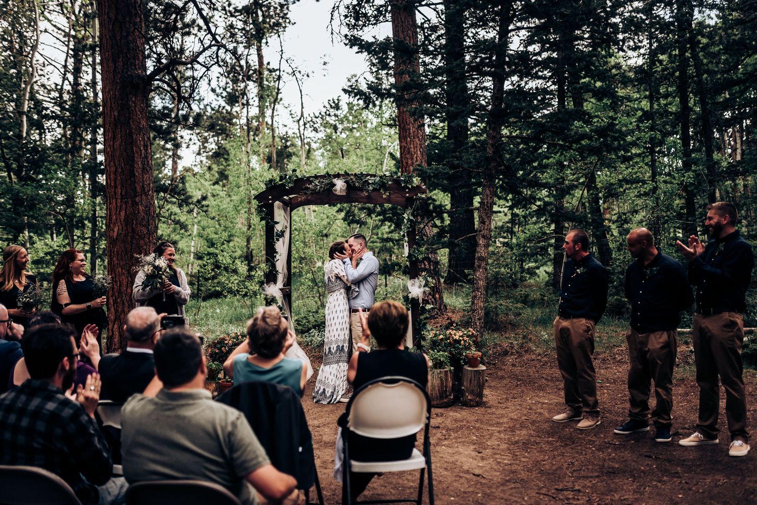 A wedding ceremony in progress beneath a flower-adorned arch surrounded by tall pine trees.