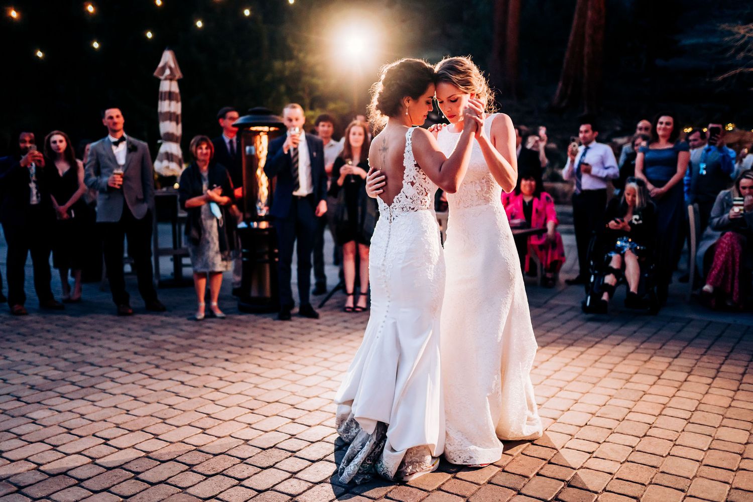 A romantic first dance at an outdoor evening wedding reception with guests watching on a brick patio under glowing lights.