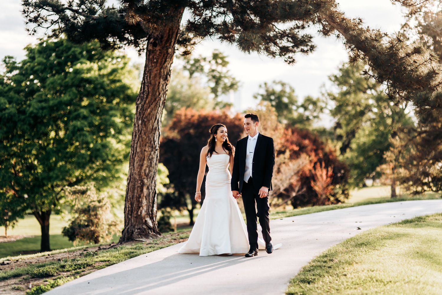 Newlyweds walking along curved path under pine trees during outdoor photo session.