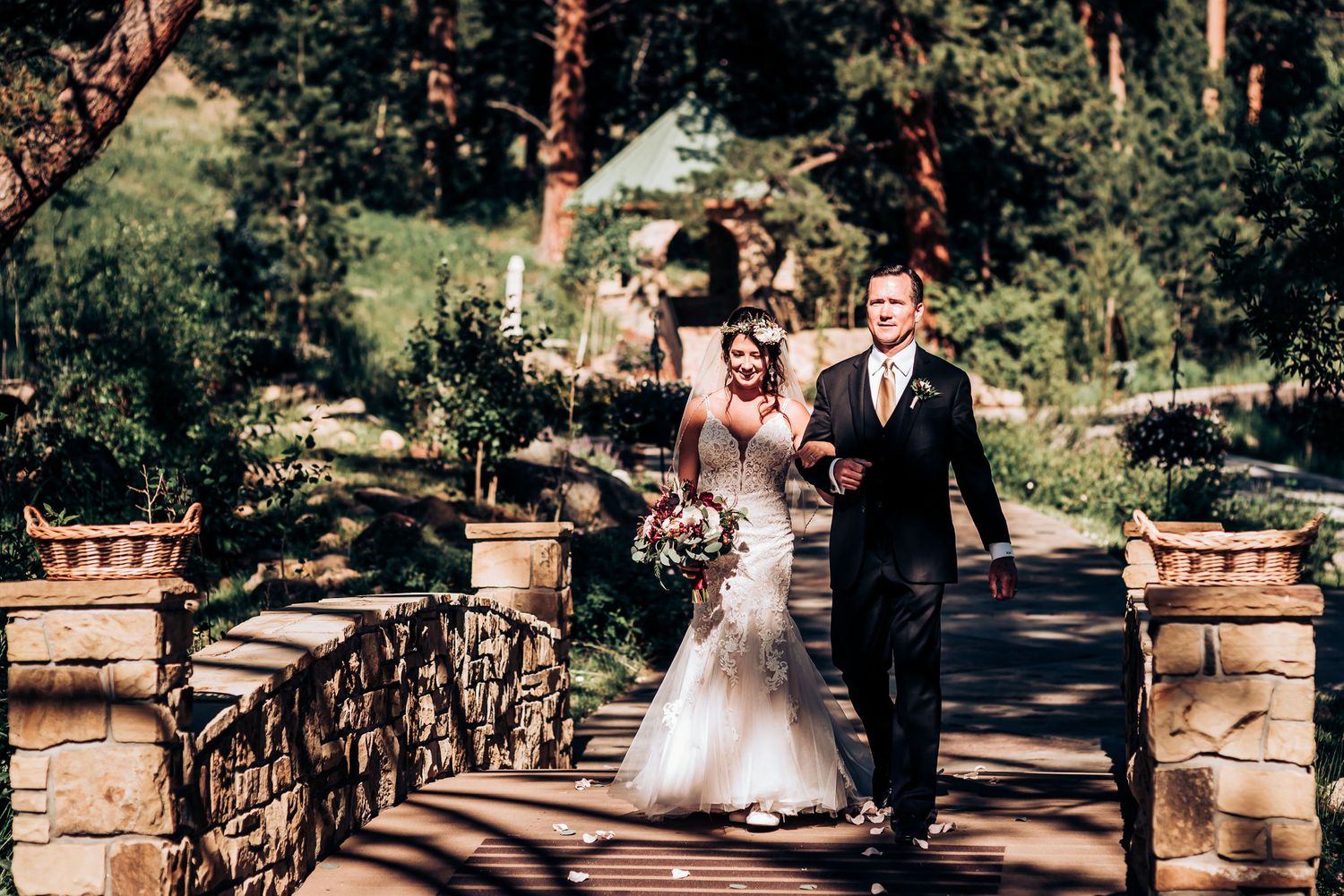 A couple walks across a decorative stone bridge during their wedding ceremony in a forest setting.