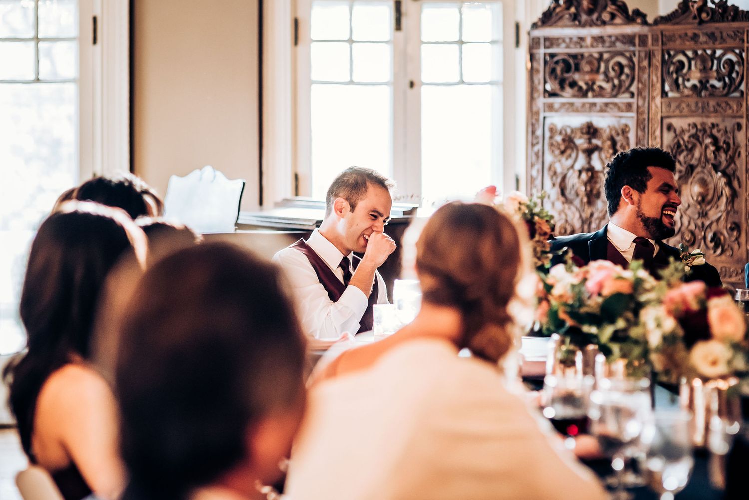 Wedding guests enjoying a reception dinner in an elegant room with ornate wooden decor and floral centerpieces.