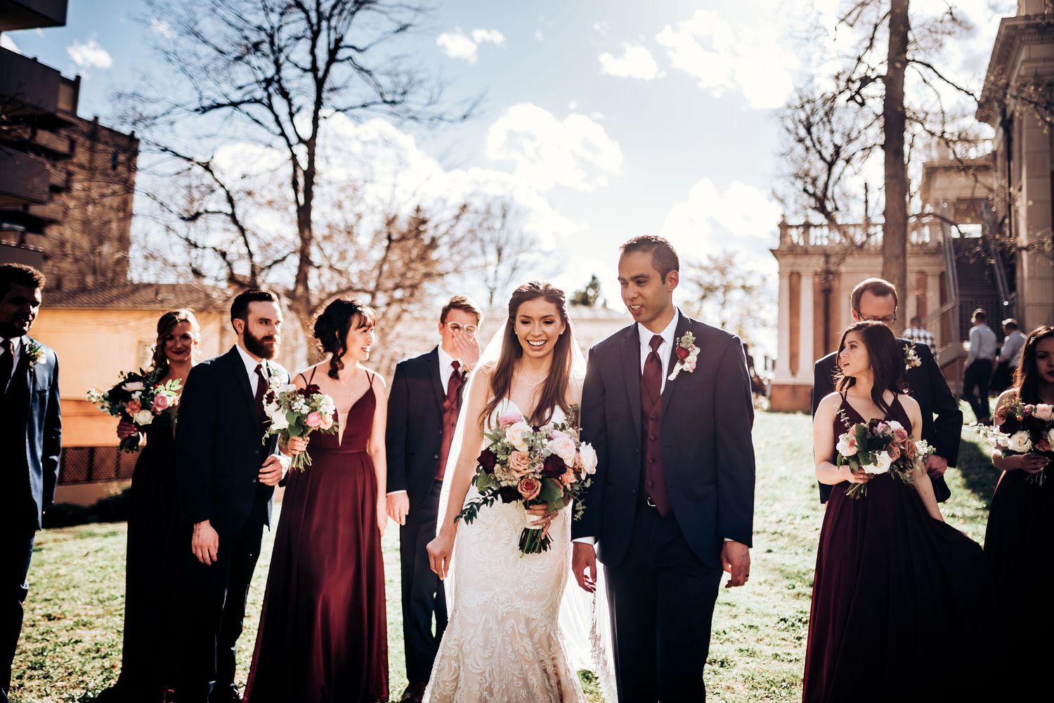 Wedding party in burgundy and black attire poses outdoors with historic architecture backdrop.