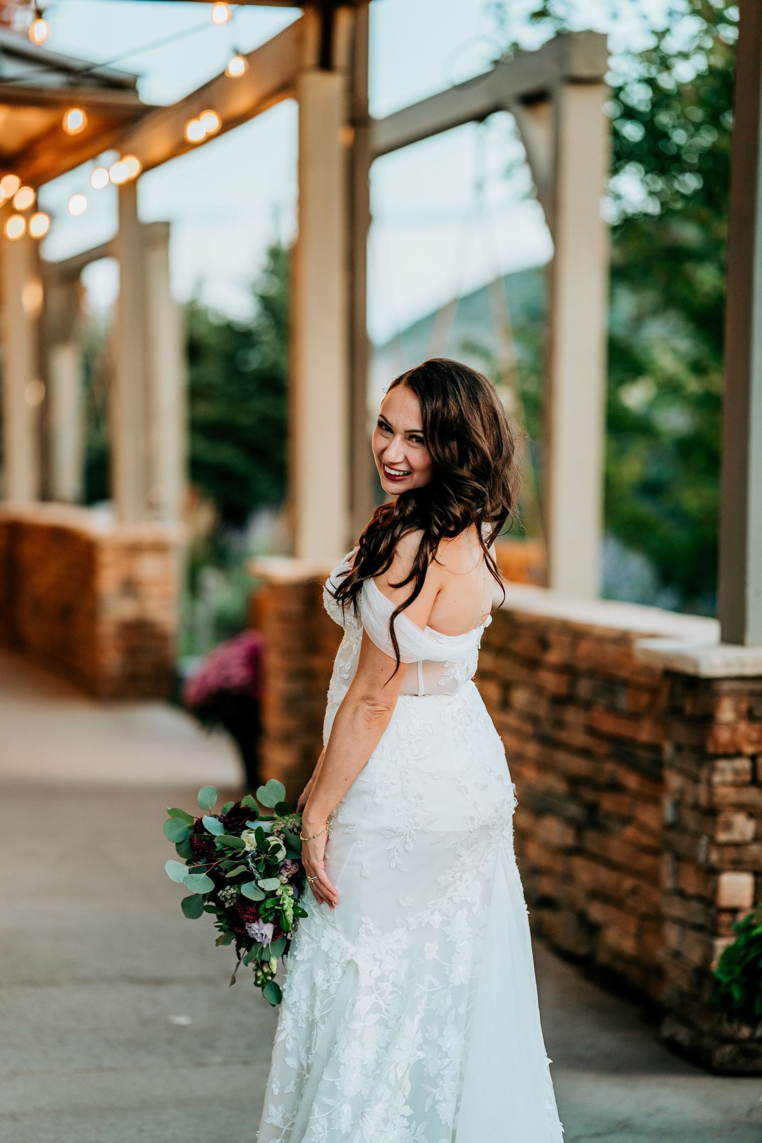 Smiling bride in off-shoulder lace wedding gown holding purple bouquet under string lights at venue.