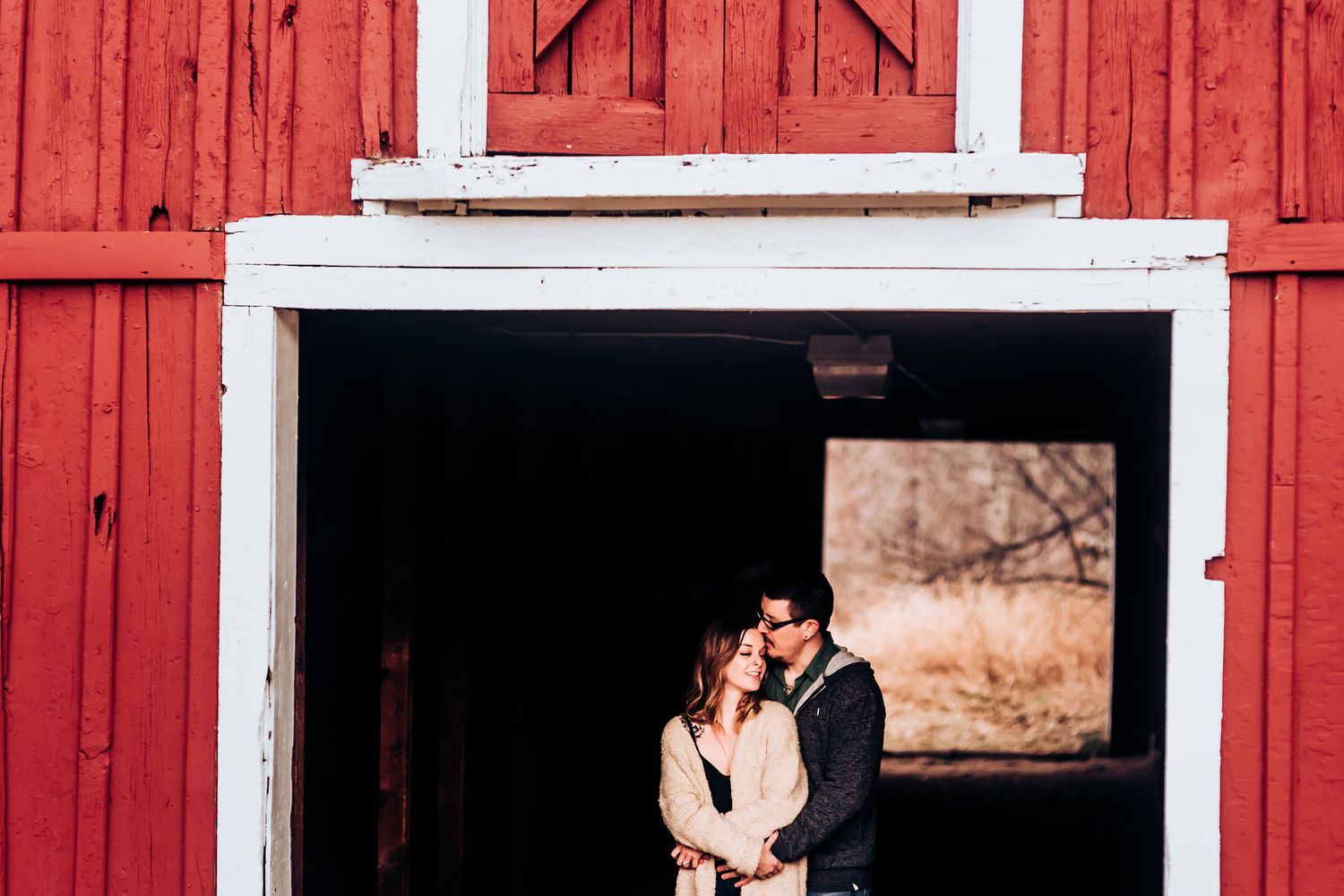A couple poses in front of a bright red barn with white trim.