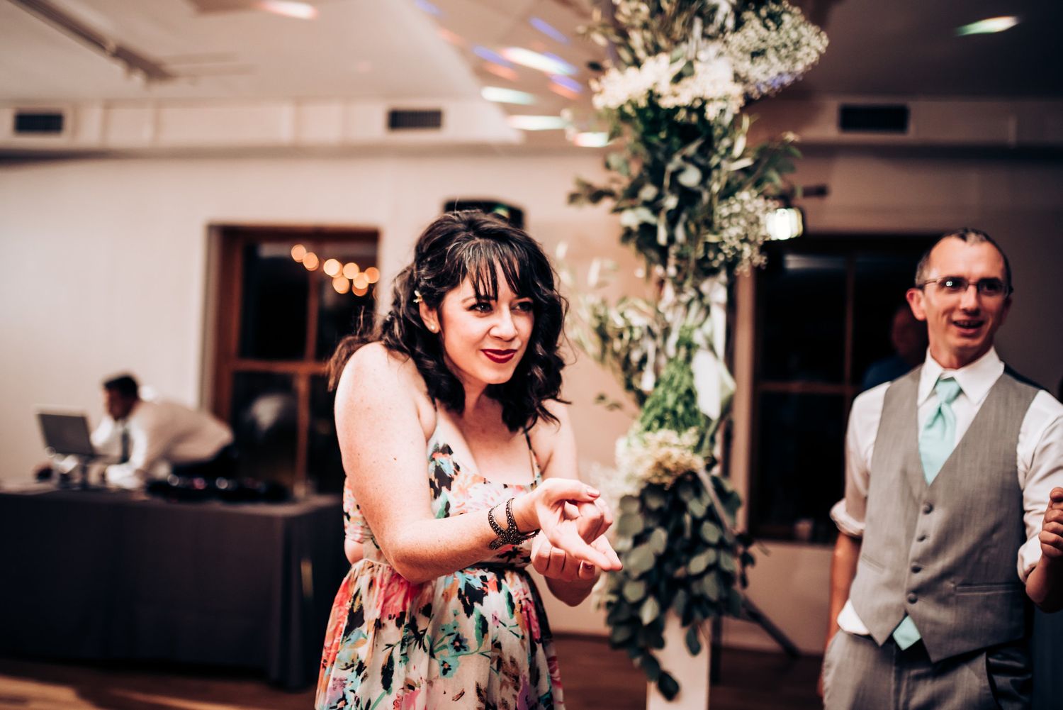 A guest in a floral dress dances joyfully at a wedding reception with decorative greenery in the background.
