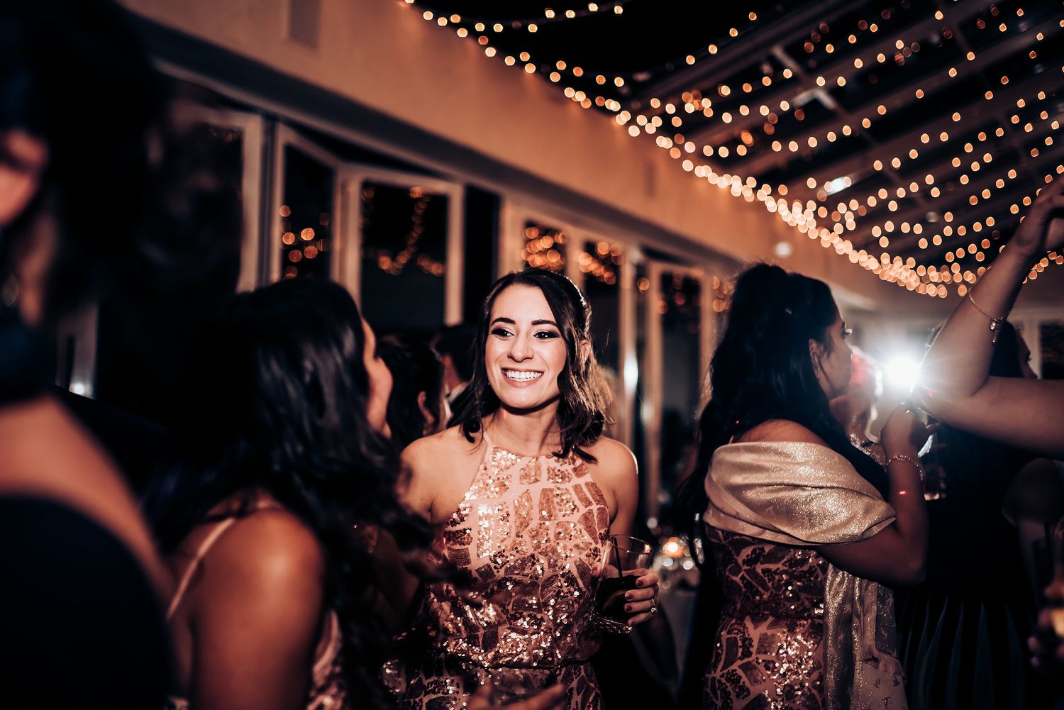 A partygoer in a sequined rose gold dress dances among others under twinkling lights.