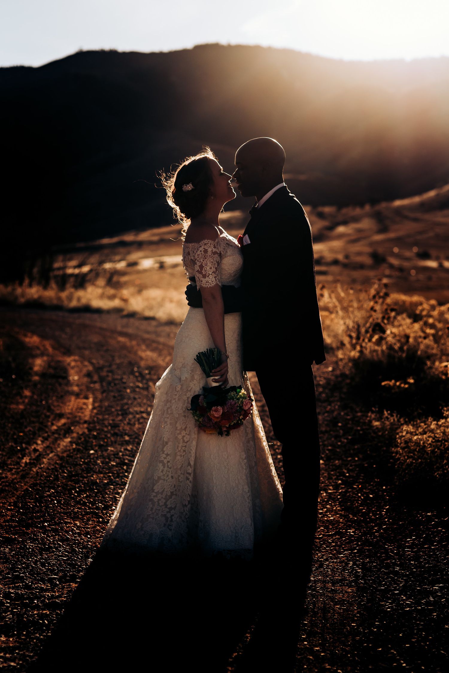 A couple in wedding attire shares an intimate moment against rolling hills during sunset.